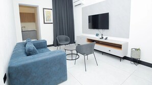 A modern living room with a blue sofa, a grey armchair, a small round table, a flat-screen TV mounted on the wall, and a kitchenette in the background. - Noble Nook Boutique Hotel (Abuja)