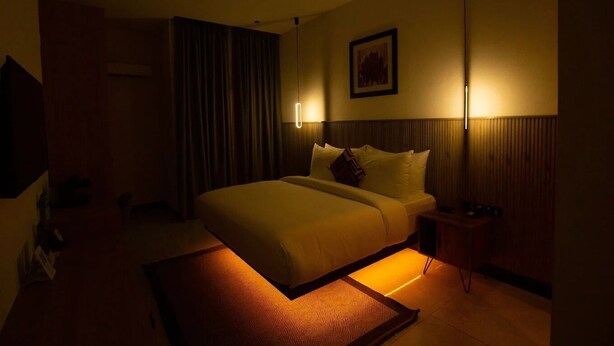A dimly lit hotel room with a large bed, a bedside table, a wall-mounted lamp, and a framed picture. - Noble Nook Boutique Hotel (Abuja)