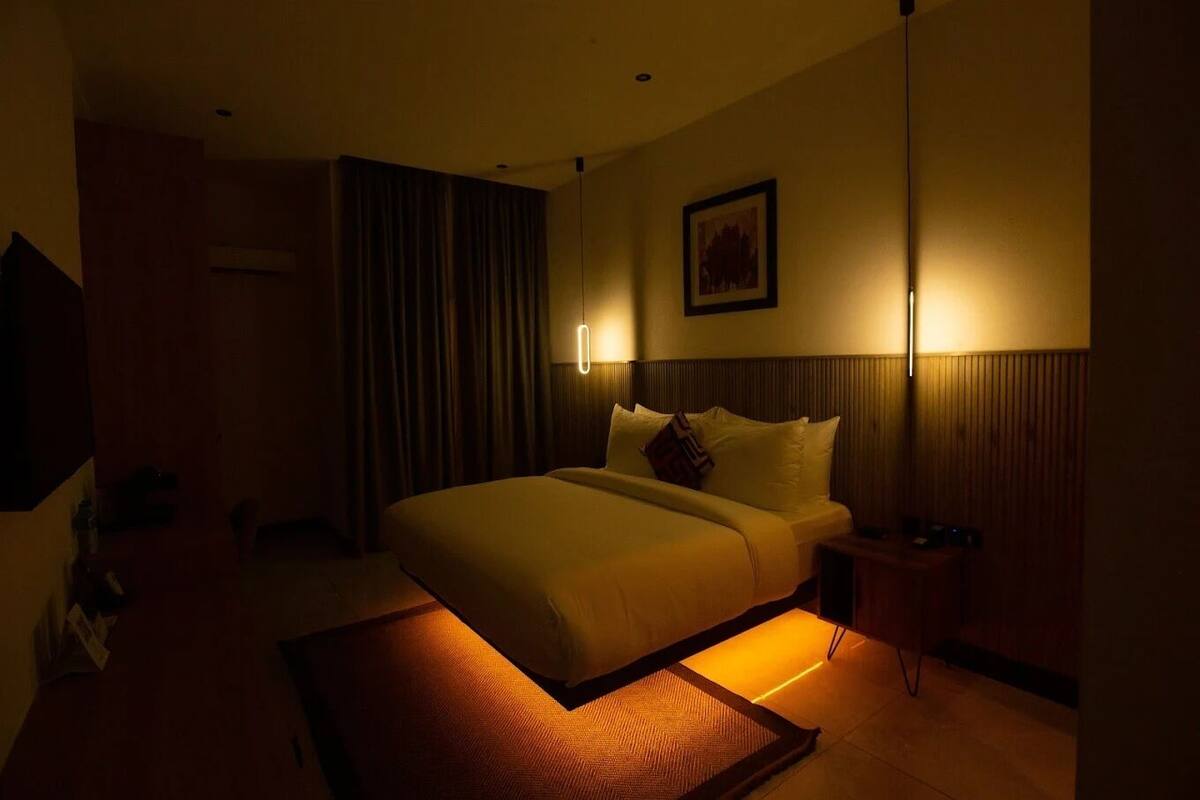 A dimly lit hotel room with a large bed, a bedside table, a wall-mounted lamp, and a framed picture.