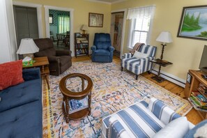 A living room with a blue sofa, a striped armchair, a blue armchair, a wooden side table, a floor lamp, and a bookshelf. - Hillside Respite by Sea Maine (Boothbay Harbor)