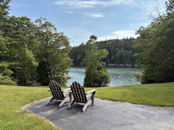 Two Adirondack chairs facing a lake surrounded by trees.