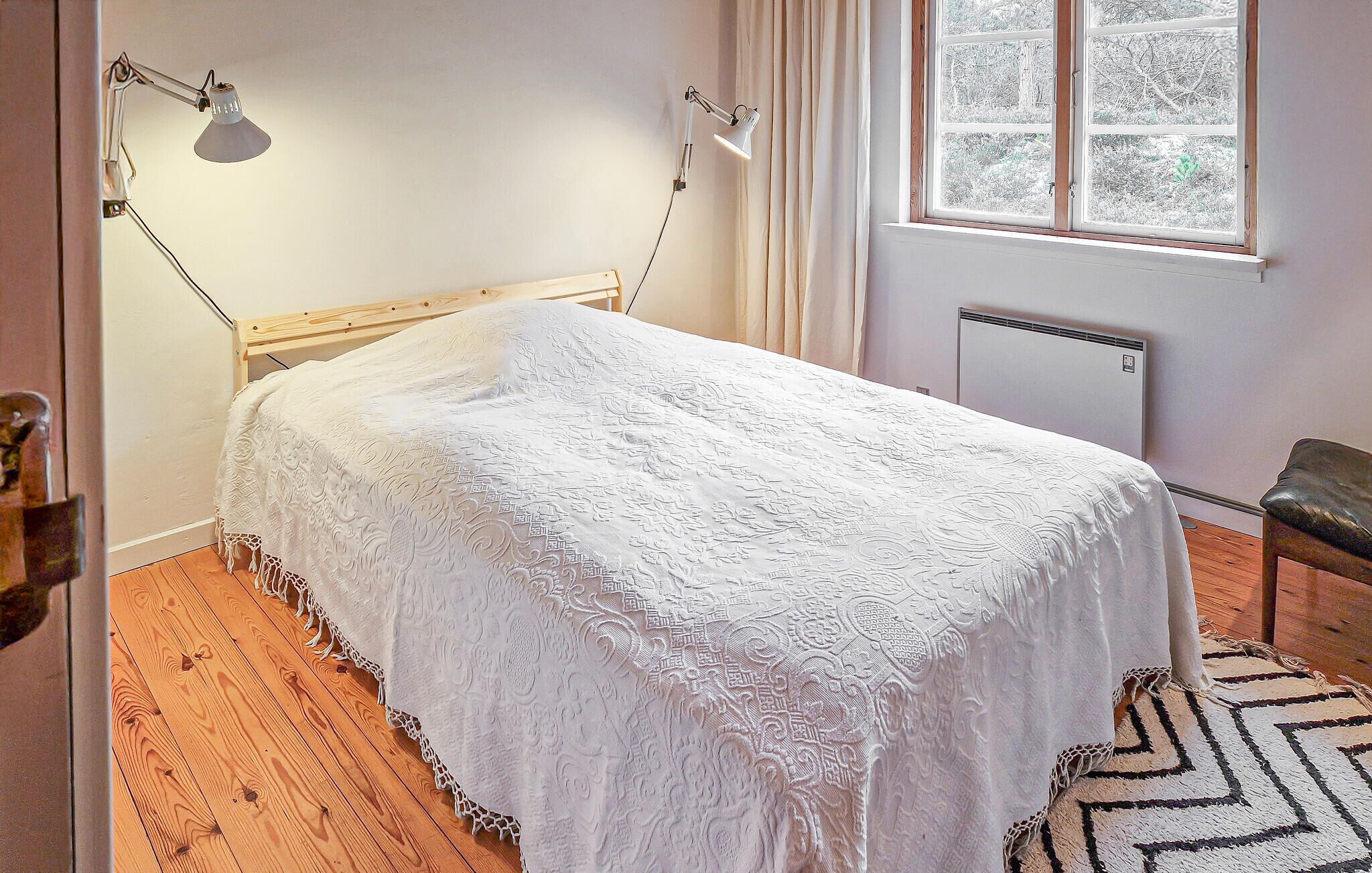 Quarto com cama de madeira, um edredom branco, duas luminárias de cabeceira, uma janela com cortinas e um radiador.