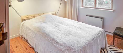 Quarto com cama de madeira, um edredom branco, duas luminárias de cabeceira, uma janela com cortinas e um radiador.