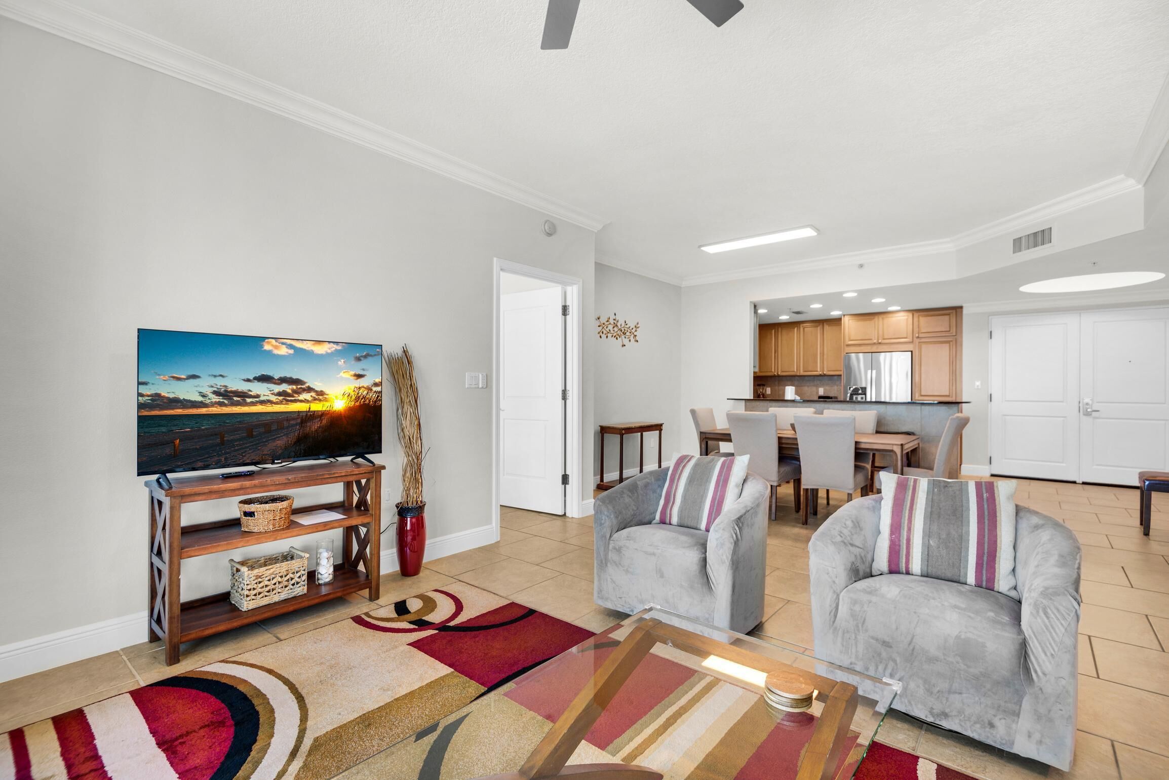 A modern living room with a flat-screen TV, two grey armchairs, a coffee table, and a kitchen in the background.