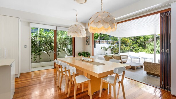 A spacious dining area with a wooden table and chairs, large windows, and a view of greenery outside. - The Shore Thing 1 Peregian Beach (PEREGIAN BEACH)