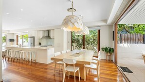 A modern kitchen with white cabinetry, a central island, and a dining area with a large table and chairs. - The Shore Thing 1 Peregian Beach (PEREGIAN BEACH)