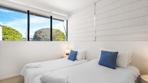 A hotel room with two beds, a window with blinds, a bedside lamp, and a view of greenery outside. - Villa Vista 1 Peregian Beach (Peregian Beach)
