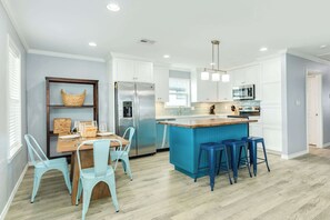 A modern kitchen with white cabinets, a stainless steel refrigerator, a blue island with stools, and a wooden dining table with chairs. - License to Chill - Your Official Permit to Relax! (Surfside Beach)