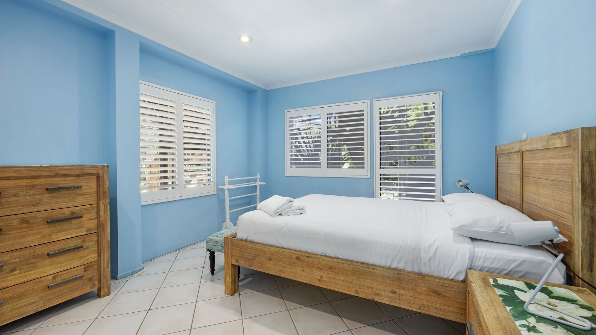 A bedroom with a wooden bed, white bedding, a wooden dresser, and a window with white blinds.
