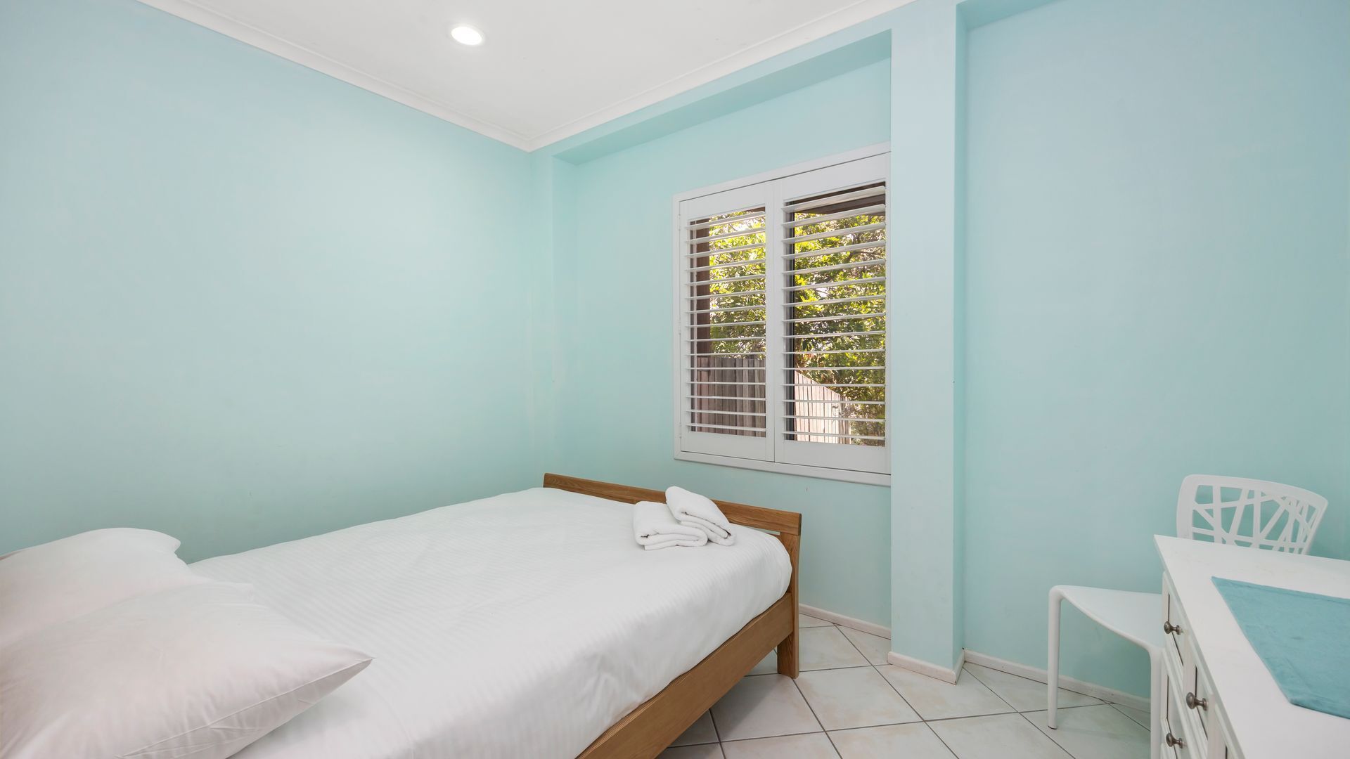 A bedroom with a single bed, a window with blinds, a chair, and a small table.
