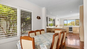 A modern kitchen with a central island, a dining area with a table and chairs, and large windows with blinds. - Piper's Bend Peregian Beach (Peregian Beach)