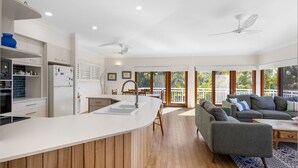 A modern kitchen with a central island, a dining area with a sofa and table, and a balcony with a view of greenery. - Notre Reve 2 Peregian Beach (PEREGIAN BEACH)