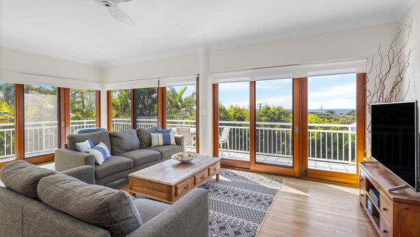 A spacious living room with a grey sofa set, a wooden coffee table, and a flat-screen TV on a wooden stand. Large sliding glass doors lead to a balcony with a white railing and a view of greenery. - Notre Reve 2 Peregian Beach (PEREGIAN BEACH)