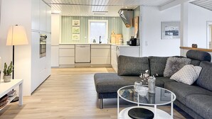 A modern living room with a grey sofa, a glass coffee table, and a lamp. A kitchen with white cabinets and a window is visible in the background. - Amazing home in Hals with house a panoramic view (Hals)