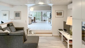 A modern living room with a grey sofa, a glass coffee table, and a TV on a white stand. There is a dining area visible through a glass door. - Amazing home in Hals with house a panoramic view (Hals)