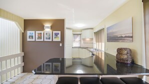 A modern kitchen with white cabinets, a black countertop, and a dining area with a glass table. - Summer Place on Shearwater Peregian Beach (PEREGIAN BEACH)