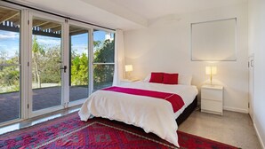 Un dormitorio con un amplio ventanal corredera que da a un balcón, una cama con ropa de cama blanca y almohadas rojas, dos mesitas de noche con lámparas y una alfombra roja con estampado.