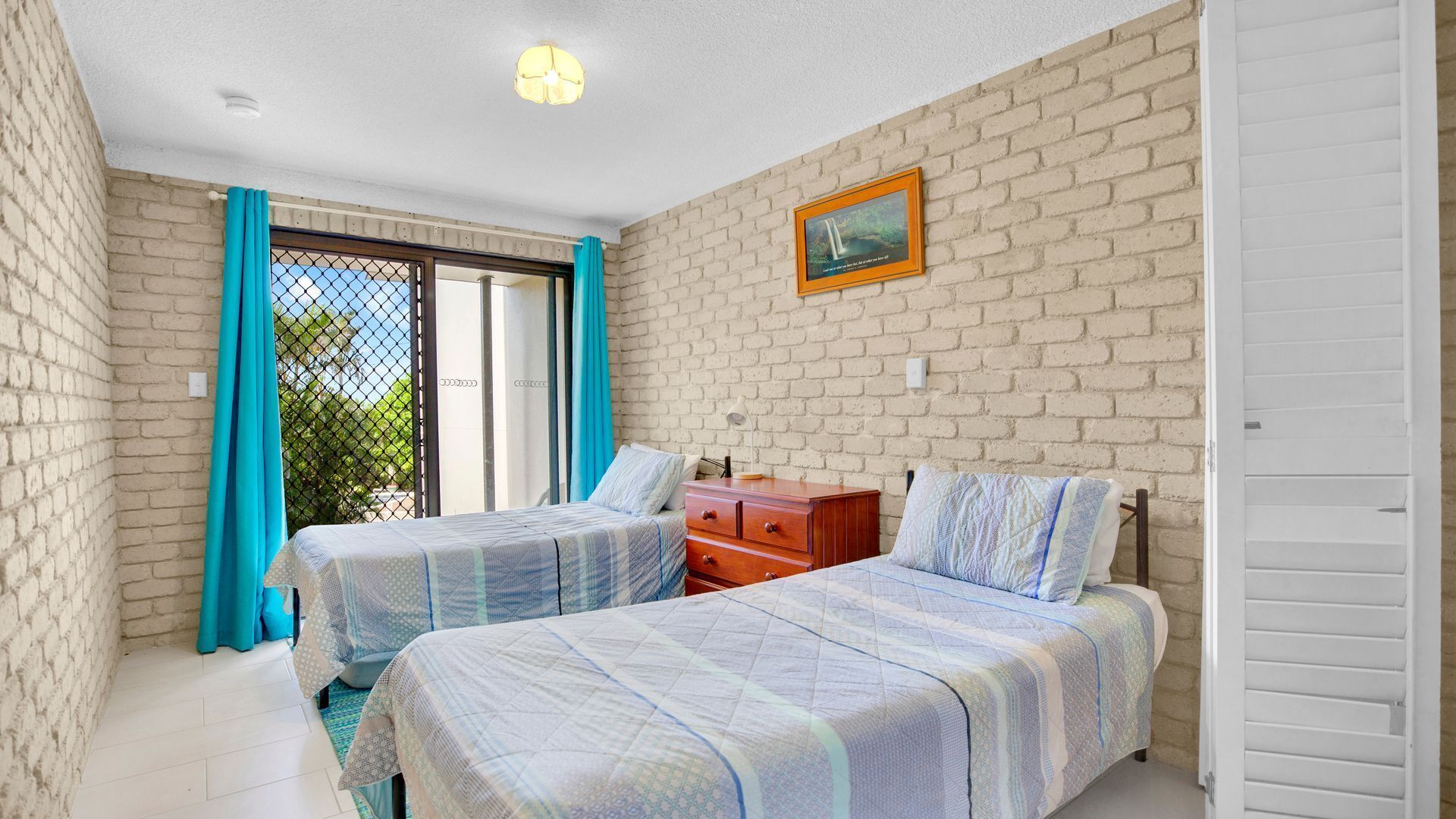 A bedroom with two beds, a wooden dresser, a white wardrobe, and a brick wall.