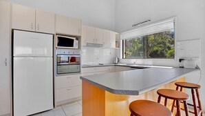 Une cuisine moderne avec des meubles blancs, un four encastré, un four à micro-ondes et une grande fenêtre offrant une vue sur la verdure environnante.
