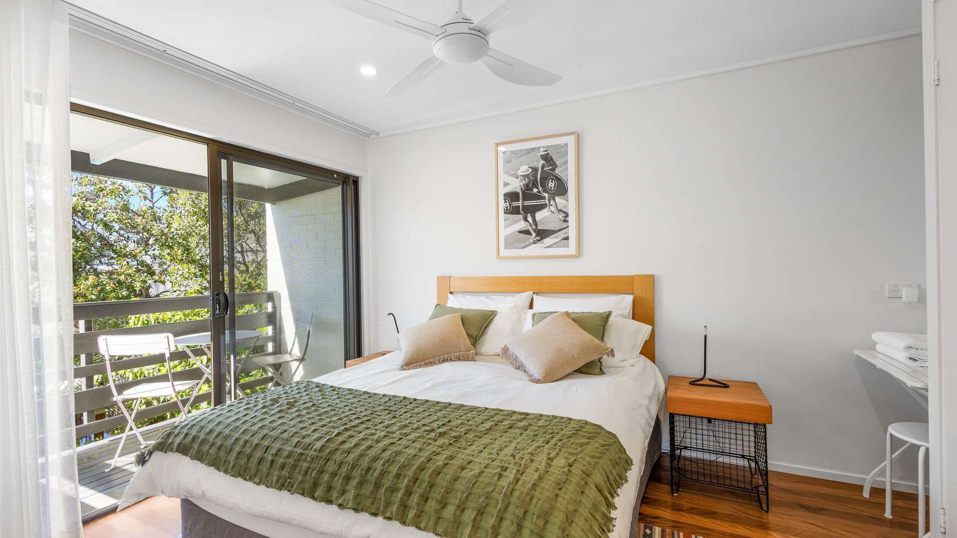 A bedroom with a bed, a ceiling fan, a framed picture, a small table, and a chair.