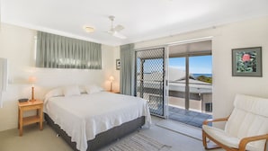 Ein Schlafzimmer mit Bett, Nachttischen, einem Sessel, einem Balkon mit Aussicht und einem Deckenventilator.