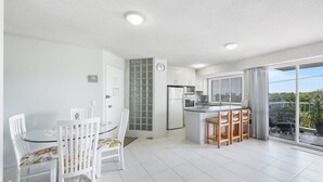Una cocina moderna con armarios blancos, un refrigerador y un microondas. Hay un comedor con una mesa de vidrio y sillas blancas. La habitación tiene un balcón con vistas a zonas verdes.