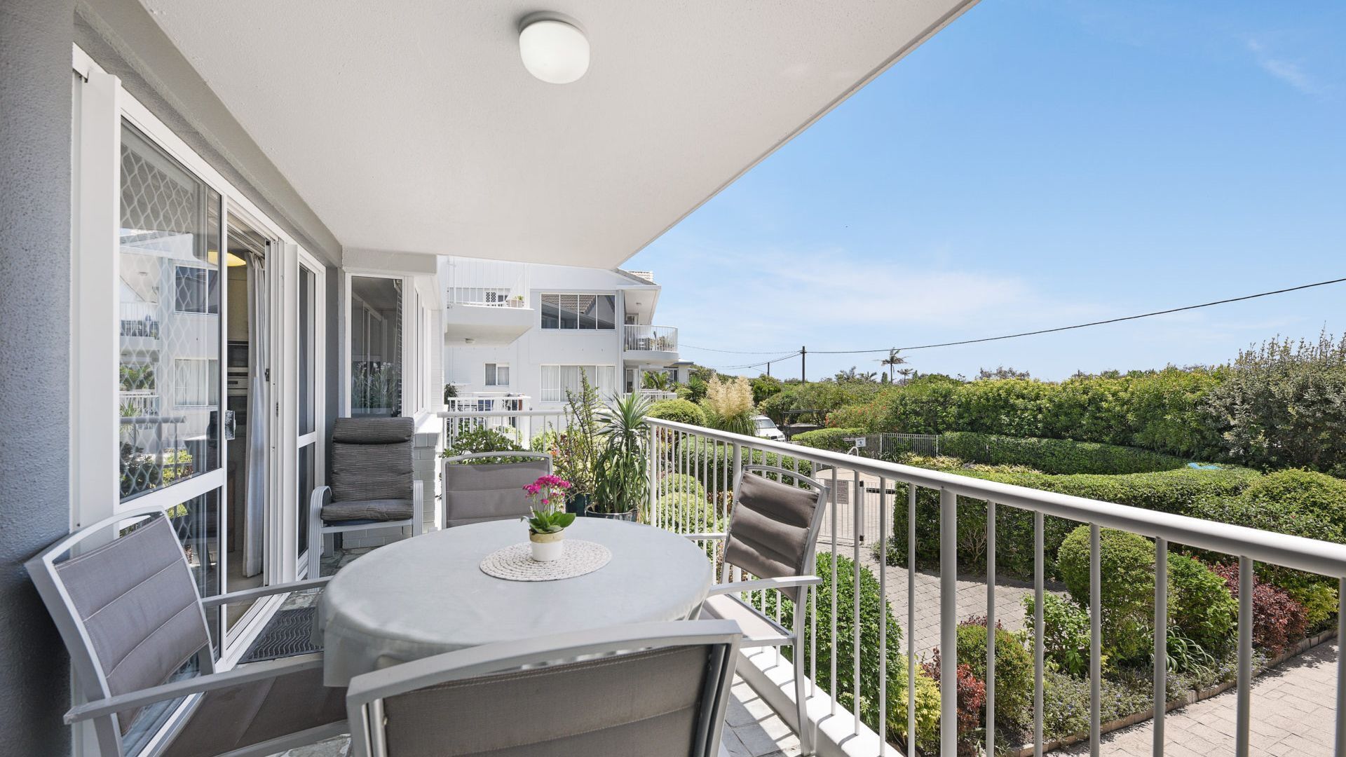 A balcony with a table and chairs, overlooking a garden and neighboring buildings.