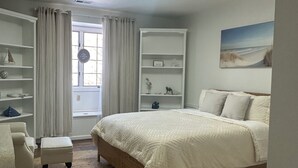 Une chambre à coucher avec un lit, une chaise, une petite table, une bibliothèque et une fenêtre avec des rideaux.