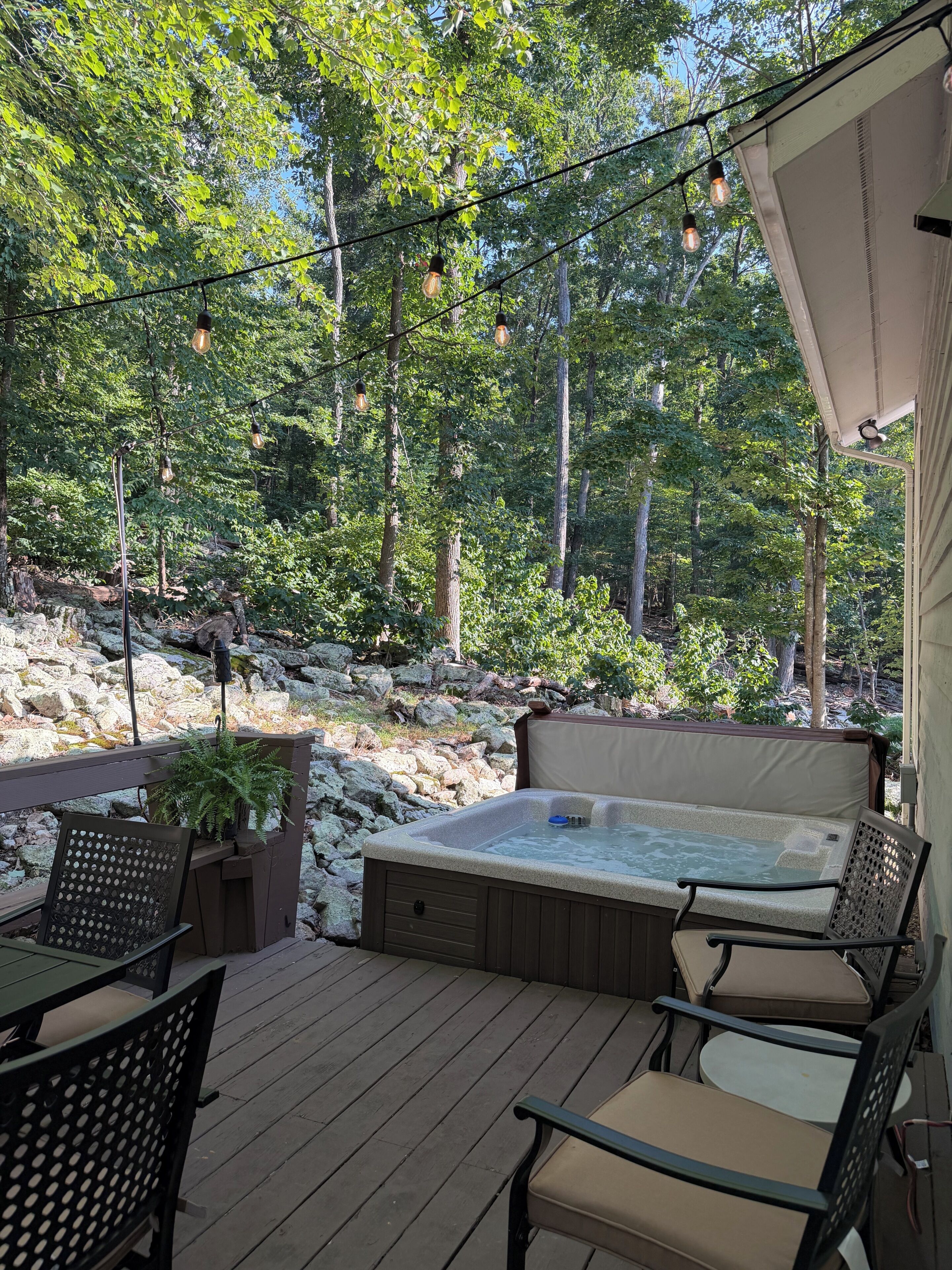 A hot tub with seating on a wooden deck, surrounded by trees and a rocky landscape.