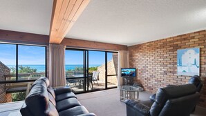 Ein geräumiger Wohnbereich mit einer großen Schiebeglasdoor zum Balkon, einem Ledersofa, einem Glastisch und einem Fernseher auf einem Standfuß.