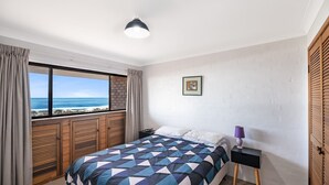 Ein Schlafzimmer mit einem großen Fenster, das Blick auf den Strand bietet, einem Bett mit einem geometrischen Muster auf der Tagesdecke, einem Nachttisch mit einer Lampe und einem gerahmten Bild an der Wand.