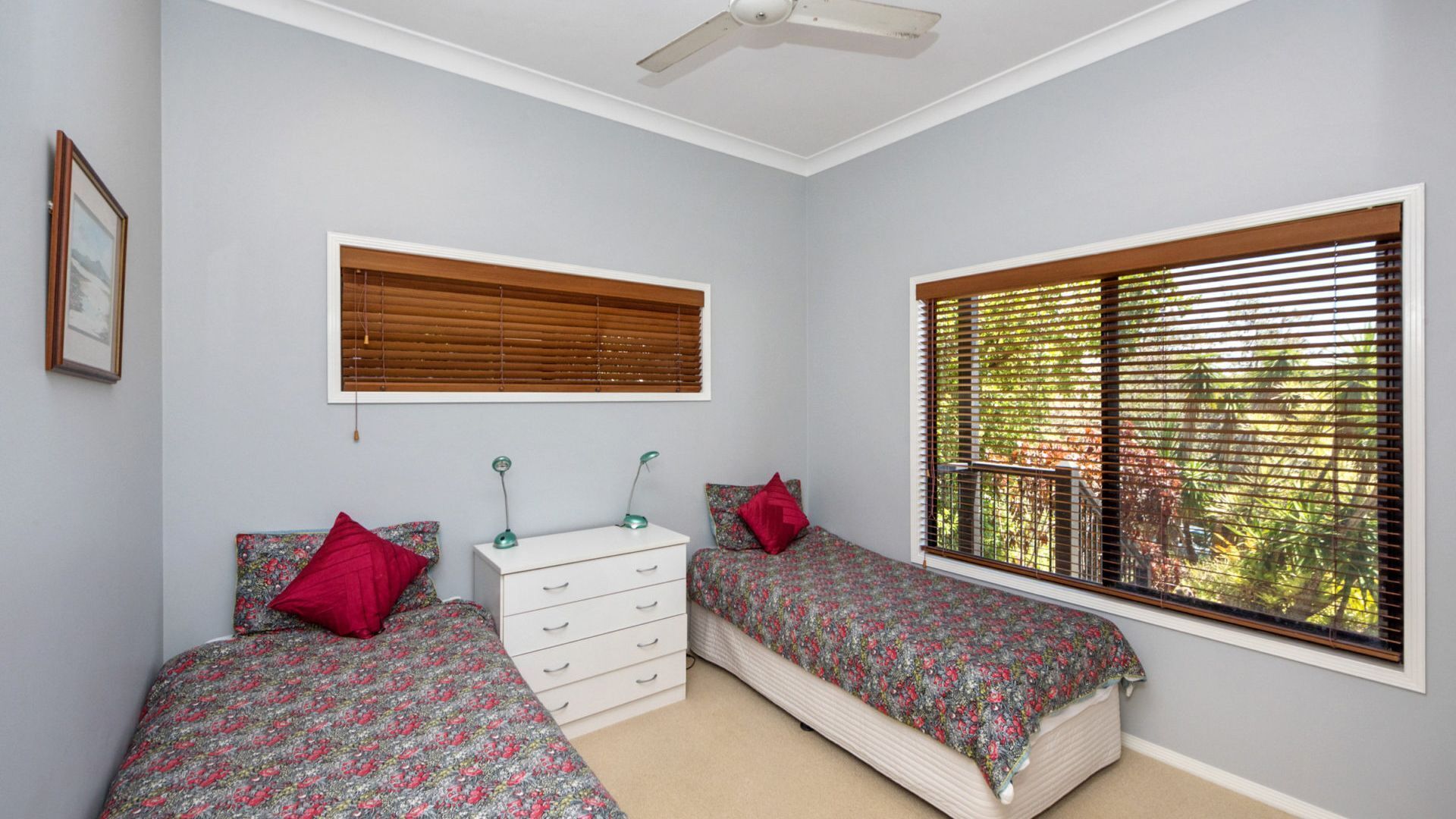 A bedroom with two single beds, a white dresser, a ceiling fan, and a window with blinds.
