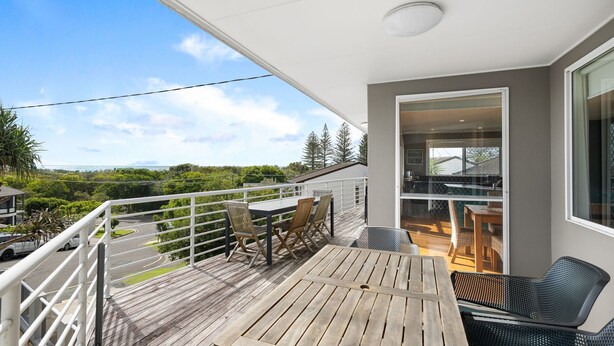 A balcony with wooden flooring, a table, and chairs, overlooking a scenic landscape with trees and a view of the sea. - Brolga Place Noosa Shire (PEREGIAN BEACH)