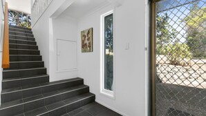 Un escalier menant à une porte dotée d’un panneau de verre, une œuvre d’art encadrée, et une vue sur un jardin à travers une porte grillagée.