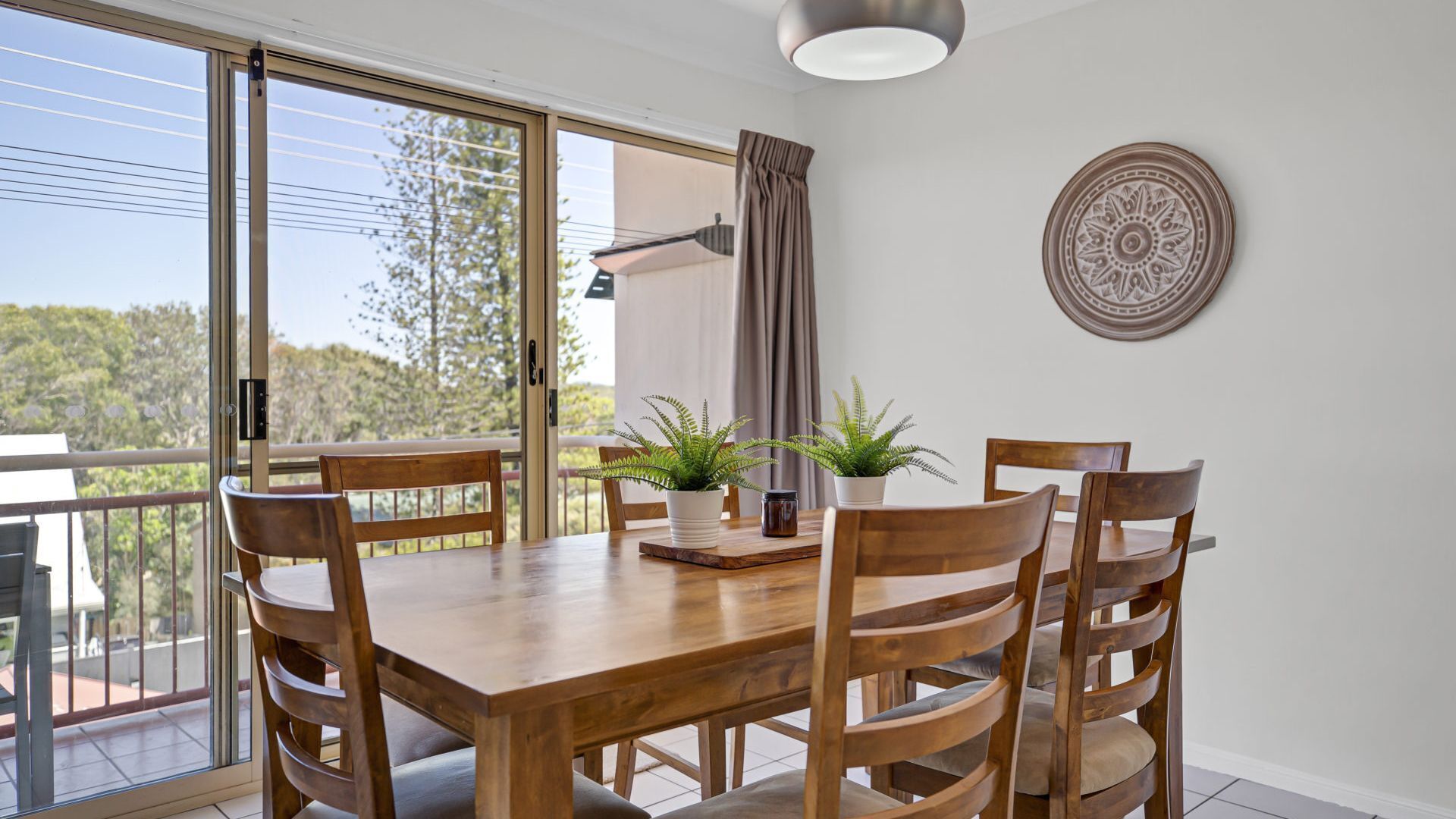 A dining area with a wooden table and chairs, two potted plants, a decorative plate on the wall, and a view of trees through a sliding glass door.