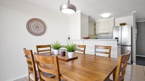 A dining area with a wooden table and chairs, a decorative plate on the wall, and a kitchen in the background. - Vaai Moana 5 Peregian Beach (PEREGIAN BEACH)