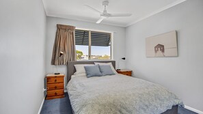 A bedroom with a bed, wooden nightstands, a ceiling fan, a window with blinds, and a painting on the wall. - Vaai Moana 5 Peregian Beach (PEREGIAN BEACH)