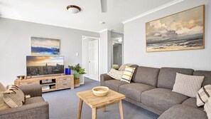 A modern living room with a grey sofa, a wooden coffee table, and a flat-screen TV on a wooden stand. - Vaai Moana 5 Peregian Beach (PEREGIAN BEACH)