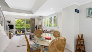 A modern living room with a glass-top dining table, wicker chairs, and a sofa. There is a large window with a view of greenery outside. - Blairgowrie 5 Peregian Beach (PEREGIAN BEACH)