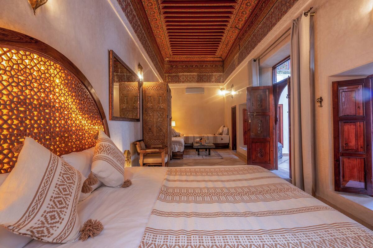 Un dormitorio amplio con una cama grande, un cabecero decorativo y vistas a una sala de estar con sofá y mesa.