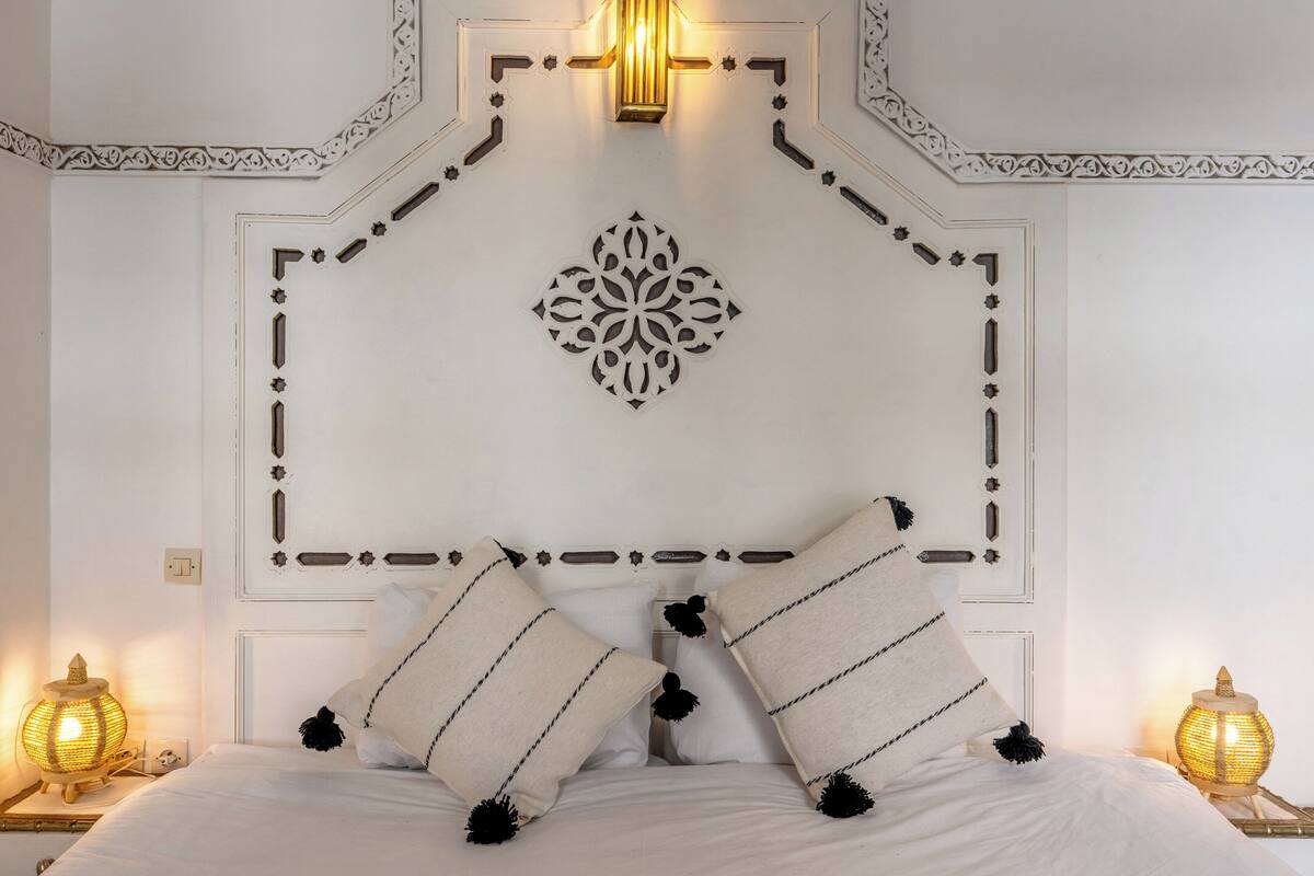 Una cama bien hecha con almohadones decorativos, una lámpara fijada a la pared y un diseño de pared simétrico.