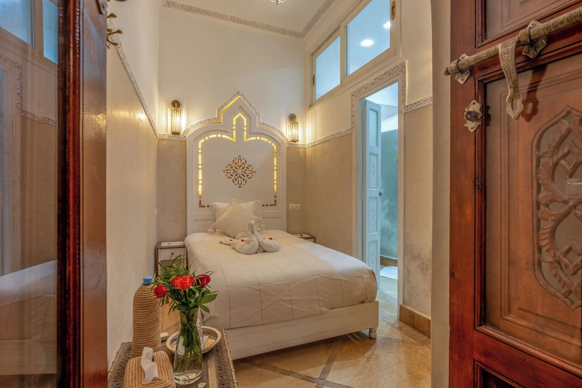 Un dormitorio bien iluminado con una cama grande, una lámpara de pared decorativa y una puerta de madera con intrincados grabados.