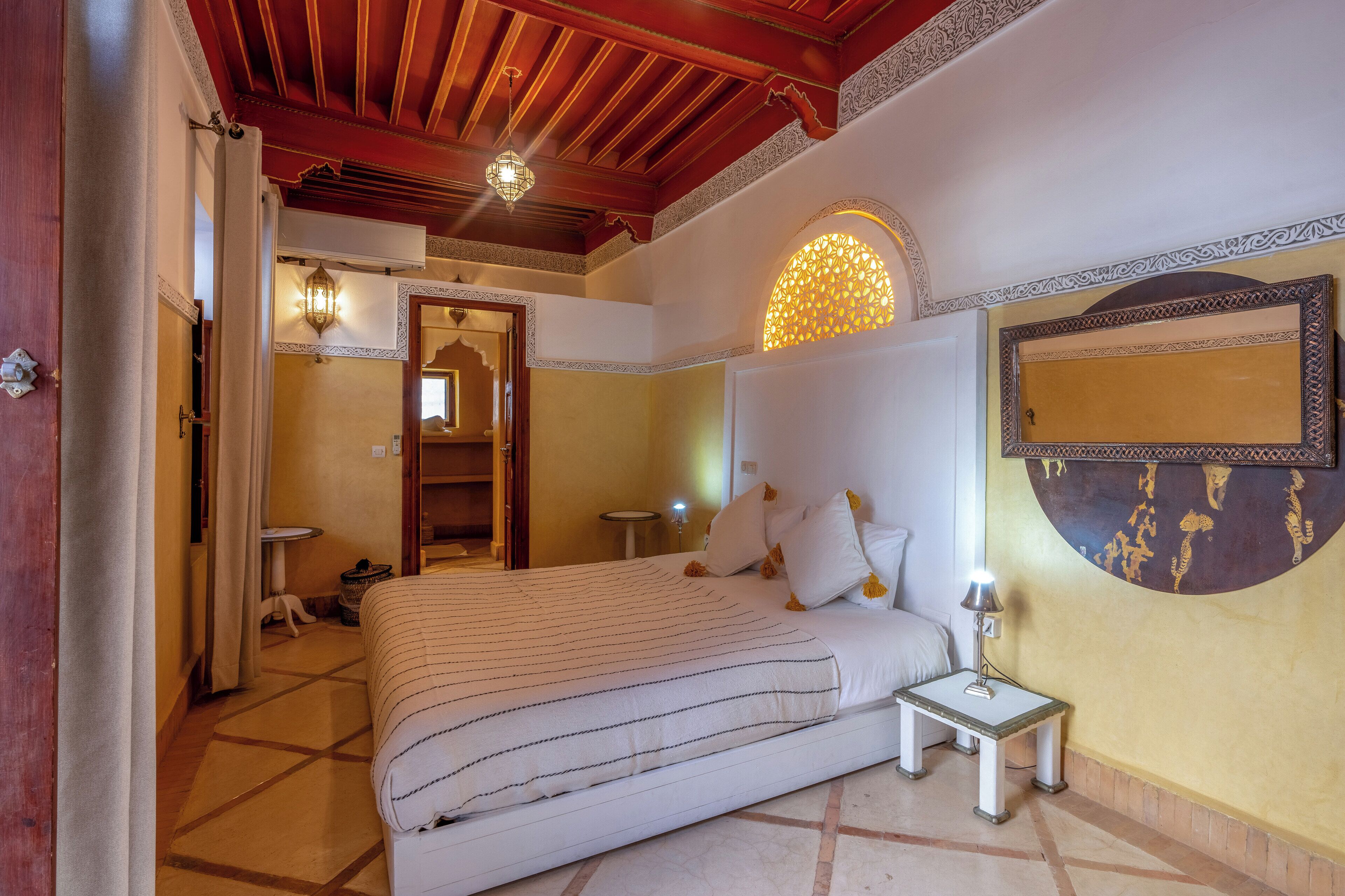A hotel room with a large bed, a bedside table with a lamp, a decorative wall piece, and a ceiling with wooden beams and a hanging light fixture.