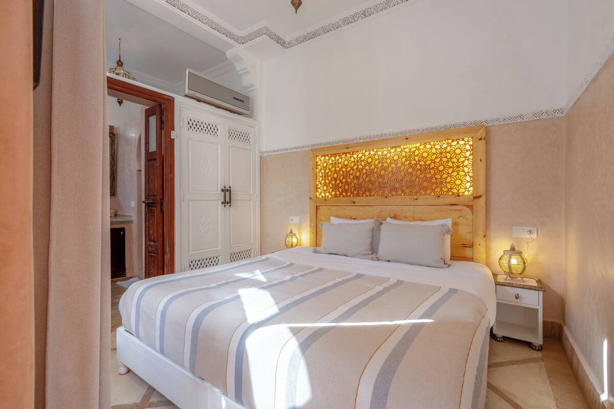 Una cama bien hecha con ropa de cama a rayas, un cabecero de madera con un panel de luz decorativo y una mesita de noche con una lámpara.