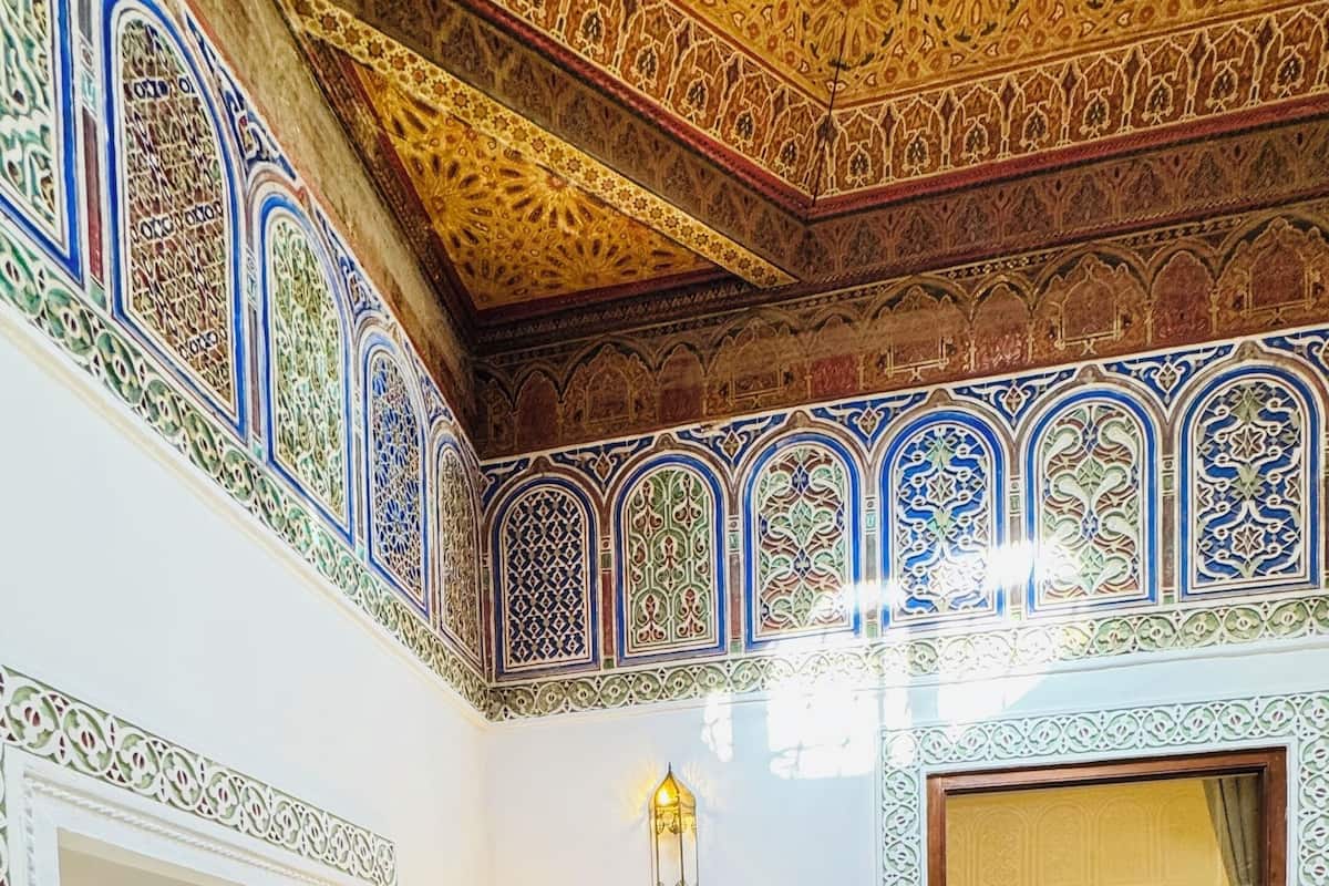 Una habitación con elaborados diseños en las paredes y el techo, un sillón verde, una pequeña mesa redonda y una cama visible a través de un arco.