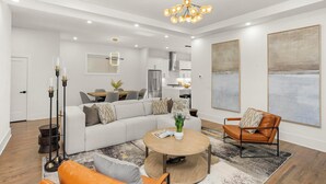 A spacious living room with a large sectional sofa, a round coffee table, and a leather armchair. There is a dining area with a table and chairs, and a kitchen in the background.