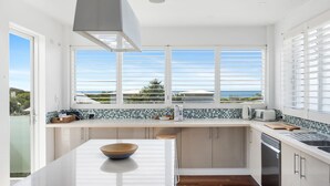 A modern kitchen with a large island, white cabinetry, and a view of the ocean through the windows. - Sea Splash Peregian Beach (PEREGIAN BEACH)