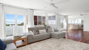 A spacious living room with a grey sectional sofa, wooden flooring, and large windows with blinds. - Sea Splash Peregian Beach (PEREGIAN BEACH)