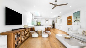 Un salon moderne avec un téléviseur à écran plat, des meubles en bois, un ventilateur de plafond et une cuisine en arrière-plan.