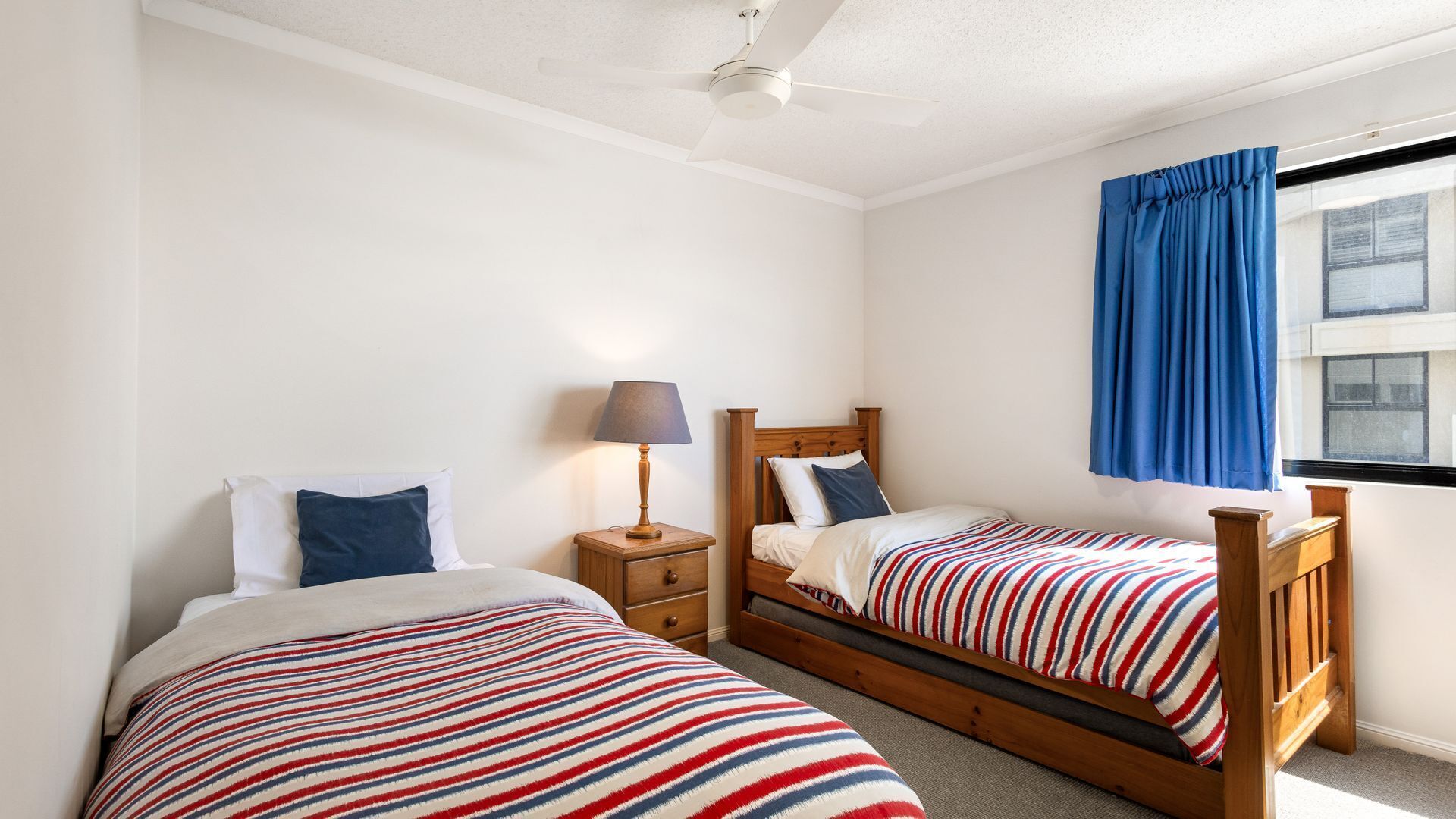 A bedroom with two single beds, a nightstand, a lamp, and a window with blue curtains.